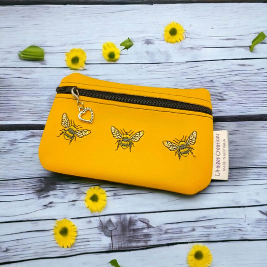 Yellow vinyl coin purse with three embroidered bees with black zip and heart zip pull (optional) sitting on wooden table with yellow daisies.