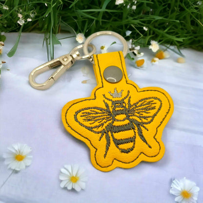 Yellow vinyl queen bee themed keychain with grey snap and silver metal keychain hardware sitting on a white table with white daisies scattered around the keychain.