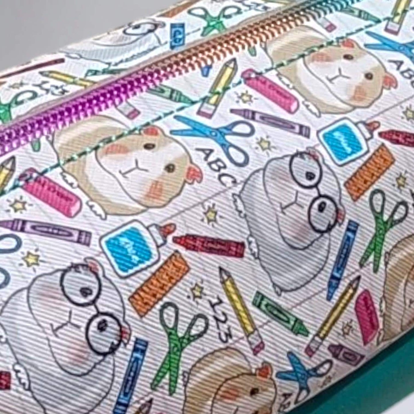 A vinyl bag with a brown and grey coloured guinea pig with scissors, crayons, pencils, glue scatted around to make up the vinyl print. The bag has a rainbow coloured zip with turquoise top stitching. The bottom of bag is made from turquoise vinyl.