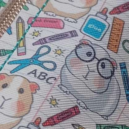 A close up of the vinyl bag with a grey coloured guinea pig with glasses. Has scissors, crayons, pencils, glue scatted around to make up the vinyl print. The bag has a rainbow coloured zip and turquoise top stitching with a rainbow heart zip pull.