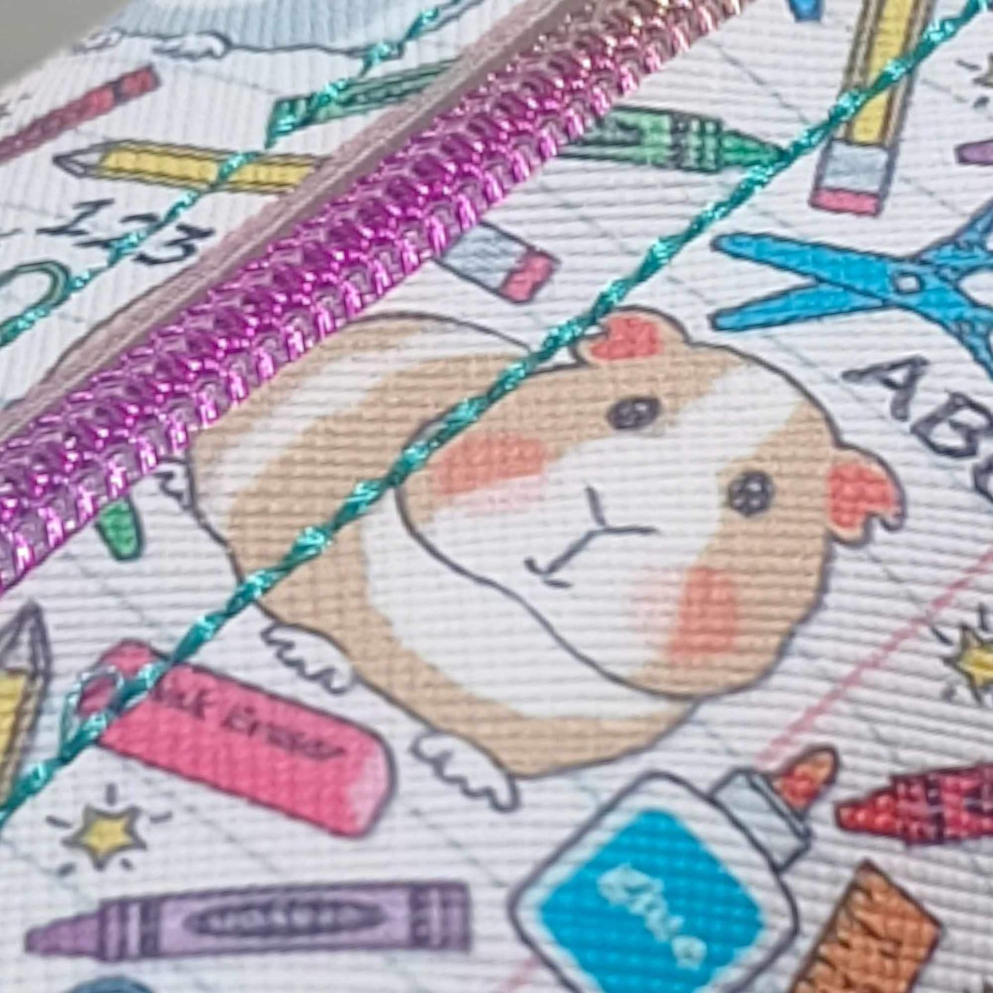 A close up of the vinyl bag with a brown coloured guinea pig with scissors, crayons, pencils, glue scatted around to make up the vinyl print. The bag has a rainbow coloured zip with turquoise top stitching.